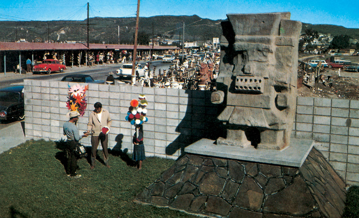 A photograph of tourists standing next to a statue of Tlaloc, the Aztec God of Rain beside a busy road.