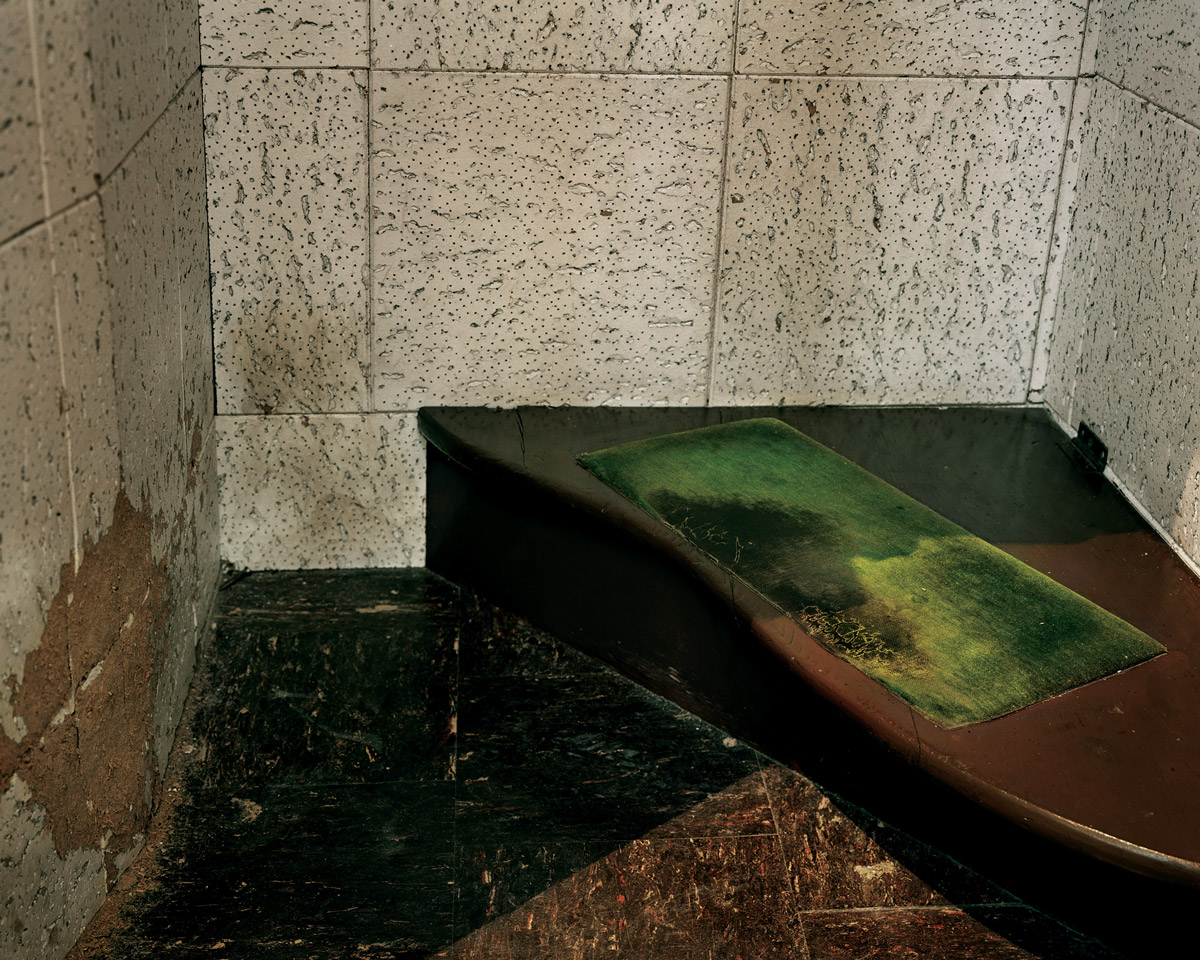 A photograph of the inside of a confessional booth with worn tile walls and a low bench, which the artist associates with Our Lady of Perpetual Help.