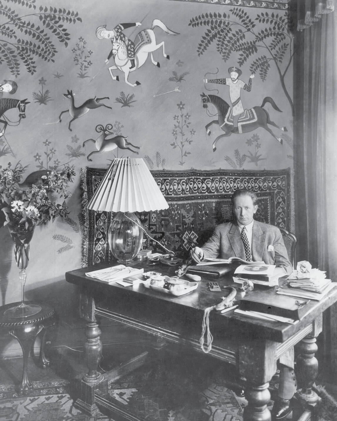 A nineteen twenty seven photograph of Murnau at a sixteenth-century Italian writing desk in his home office at Douglasstrasse twenty two. Behind him is a mural by Walter Spies of men at hunt.