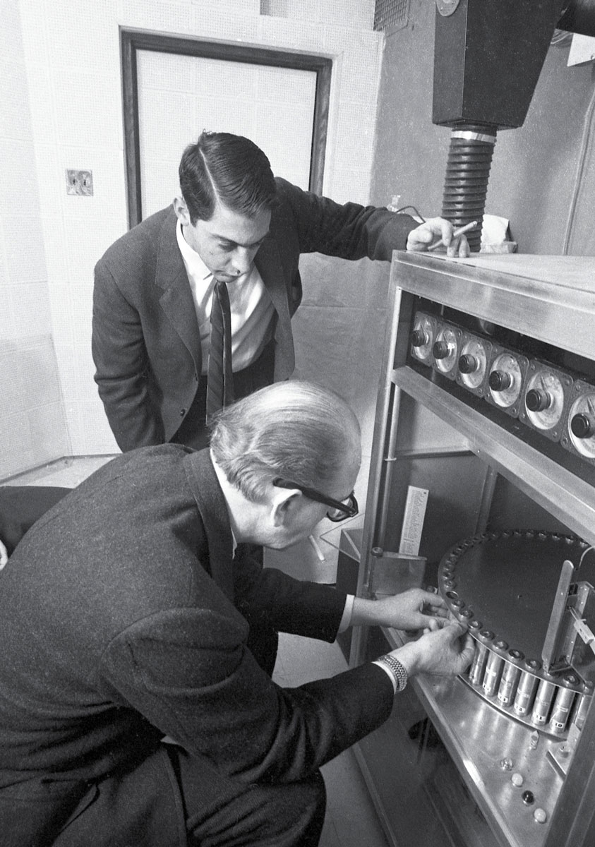 A photograph of Mike Todd Jr. observing Hans Laube fiddling with his scent energizer on the night of its world premiere. A large structure connects to smaller tubes that carry the scents to the theater above.