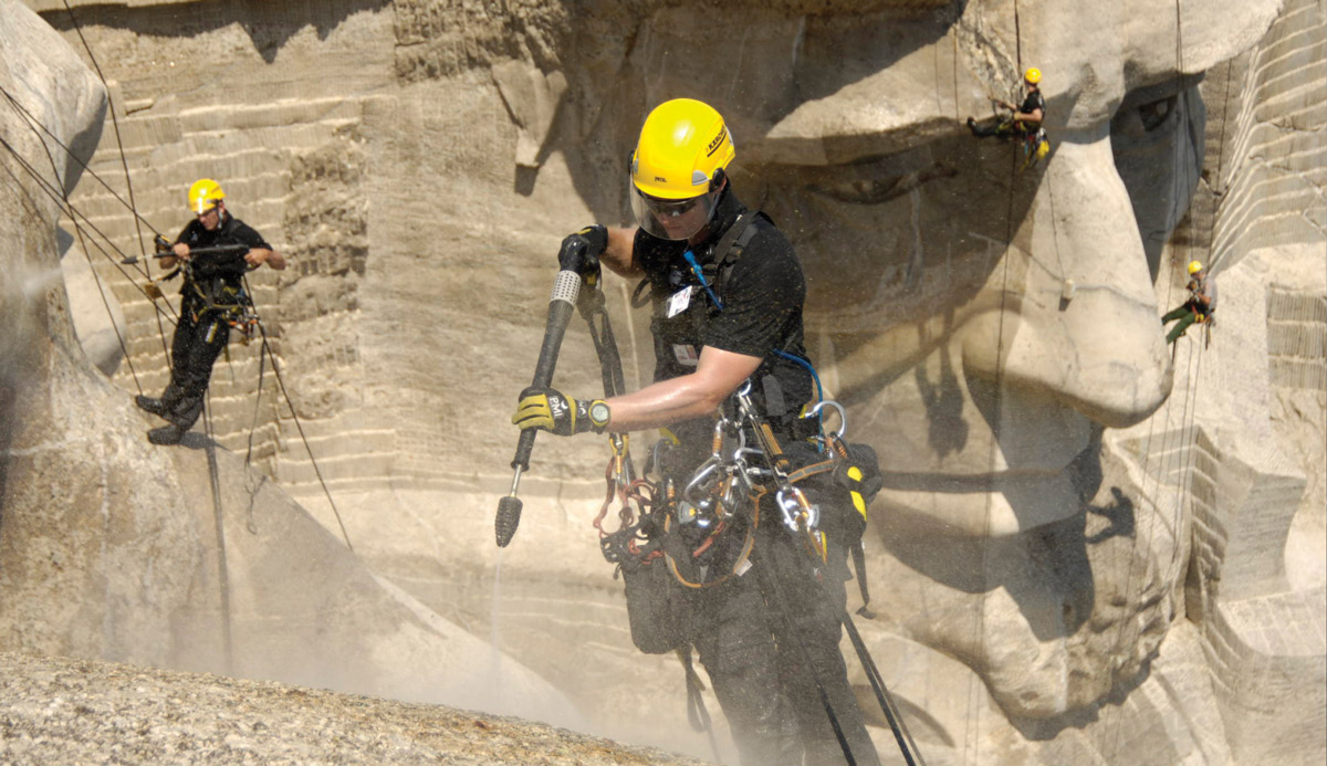 A photograph of workers tethered by suspension to the side of Mount Rushmore. The workers are drilling into various parts of the sculpture and Abraham Lincoln's face is visible in the background.