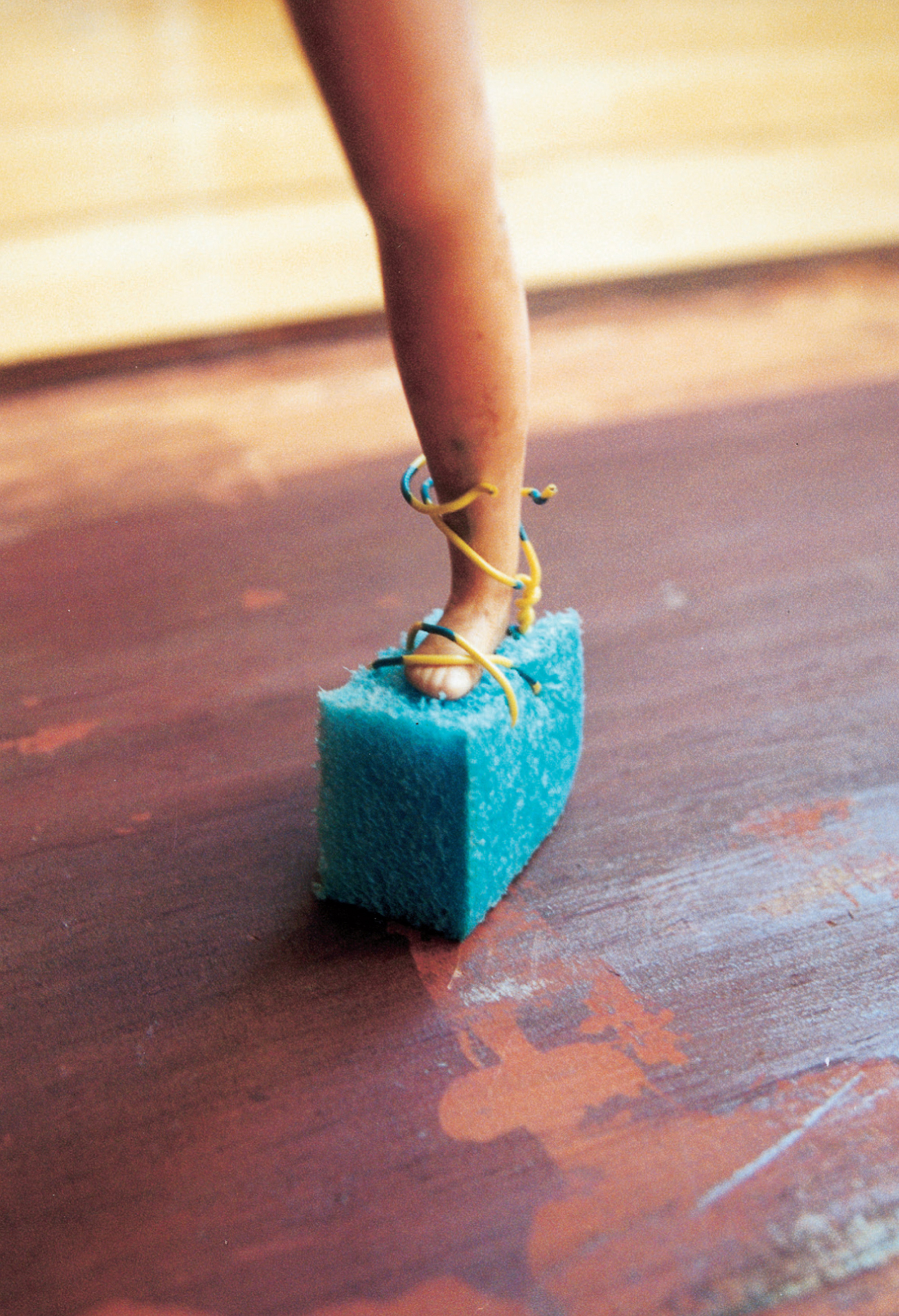 A photograph depicting a small piece of blue foam attached to a doll's foot. The caption reads: Single platform sandal of blue styrofoam with straps of yellow and blue telephone wire securing the toe with an X but loose at the ankle, up which they are possibly intended to be crisscrossed, Roman style. This artifact exemplifies the Doll Games’ strategic appropriation and ironic framing of elements of the dominant culture (in this case, popular styles of the disco era). The care put into crafting an article that would have been spurned by the Doll Games’ androgynous heroines shows the complex interplay of desire and scorn in the Doll Games’ ongoing interrogation of femininity. Just whose foot did the cobbler have in mind? Like the bewildered prince in the fairy tale, we are left with a shoe and a question.
